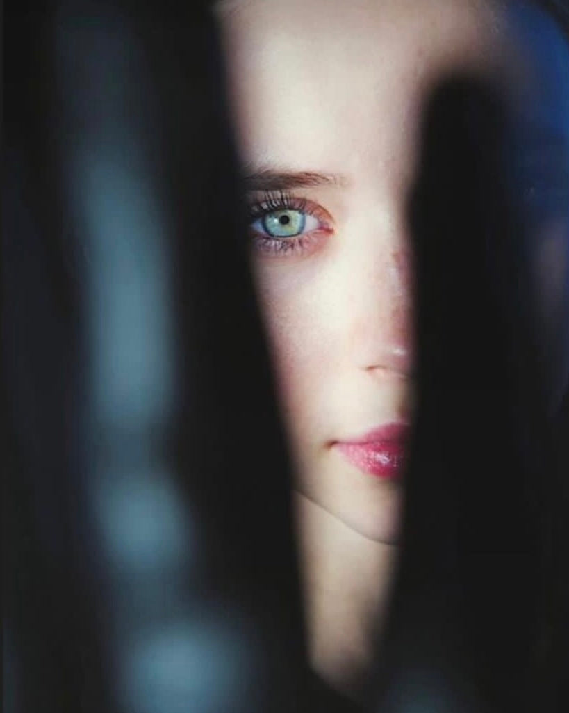 Photographie du visage d’une femme par la photographe Vanessa Moselle. Son visage est en partie caché et nous ne voyons que la moitié gauche du visage. Son œil est bleu vert, elle a un rouge à lèvres rose/rouge.