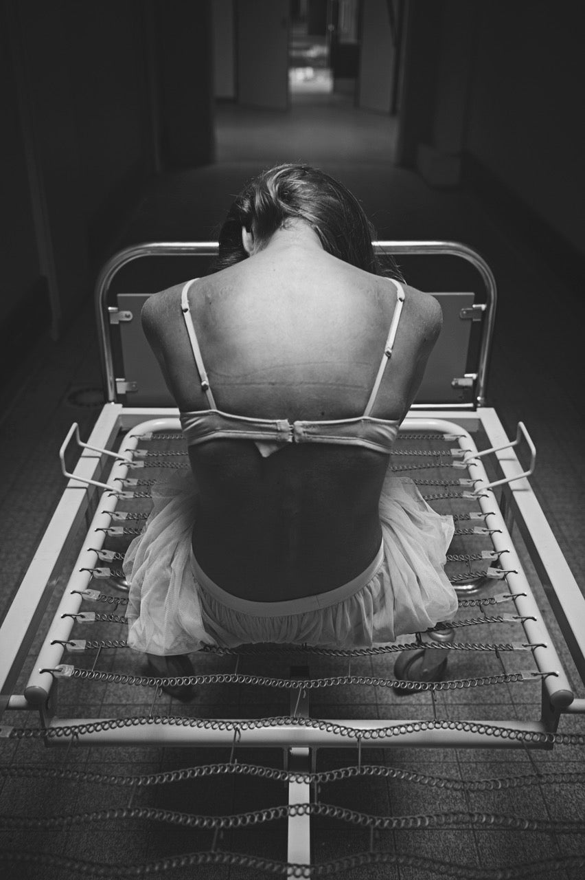 Photographie en noir et blanc réalisée par l’artiste Vanessa Moselle. Une femme est dos à nous, assise sur un lit d’hôpital sans matelas au milieu d’un couloir. Elle porte un soutient gorge blanc et une jupe à volants blanche.