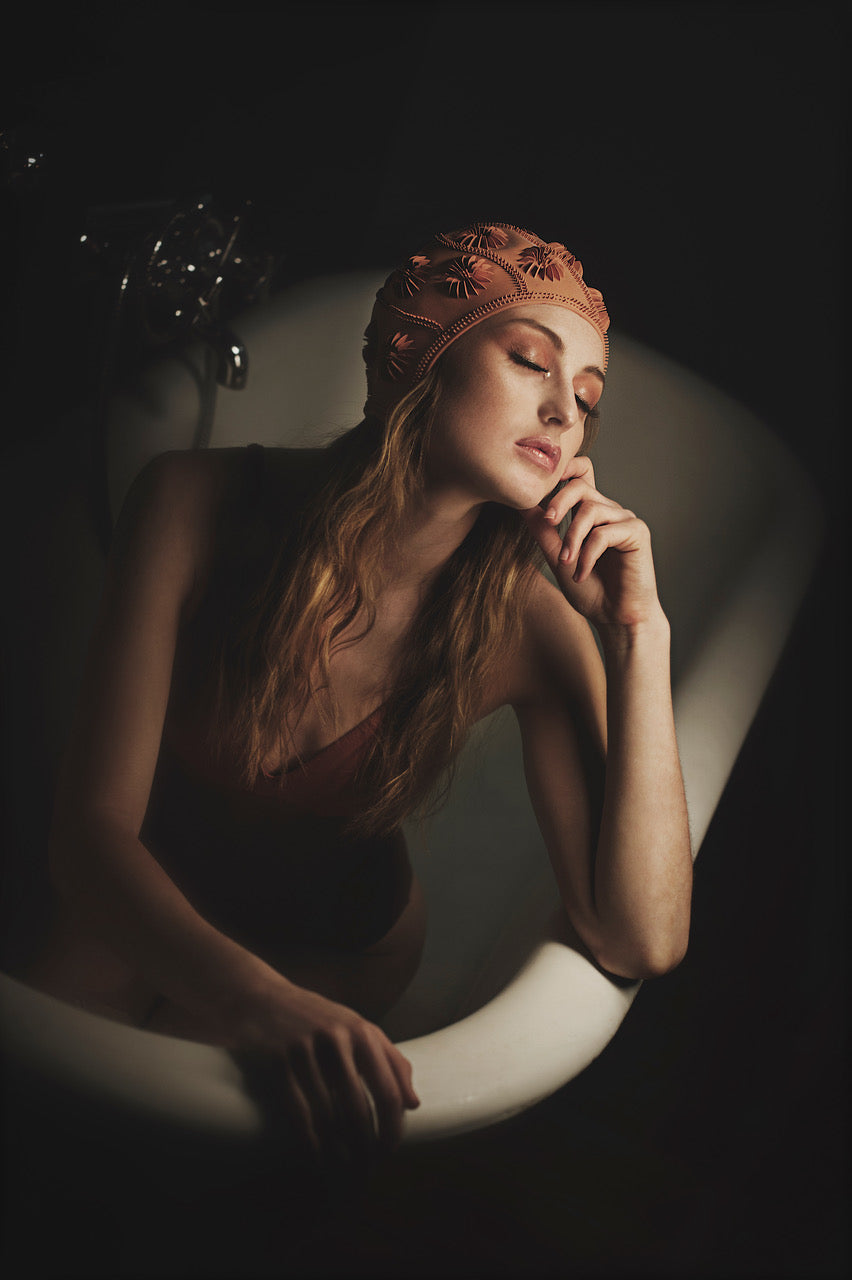 Une jeune femme se trouve assise dans une vieille baignoire. Elle a le coude posé sur le rebord de la baignoire et sa main supporte sa tête. Ses yeux sont fermés et elle a l’air pensive. Elle porte un bonnet de piscine de couleur ocre et un maillot de bain une pièce. Photographie réalisée par Vanessa Moselle.