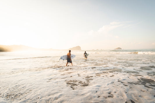 Photographie de deux surfeurs prise à Manuel Antonio au Costa Rica par l’artiste Yorick Serriere. Les surfeurs portent leur planches sous le bras et ont l’eau au niveau des genoux. La photographie est très lumineuse et le blanc de l’écume se confond avec le ciel.