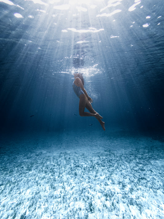 Photographie d’une femme sous l’eau prise par l’artiste Yorick Serriere en Corse en France. La femme est au centre de l’image, elle porte un maillot de bain une pièce rayé, sa tête atteint la surface. L’image est d’un bleu profond, les rayons du soleil traversent l’eau et amènent la lumière au centre de l’image juste au dessus de la femme.