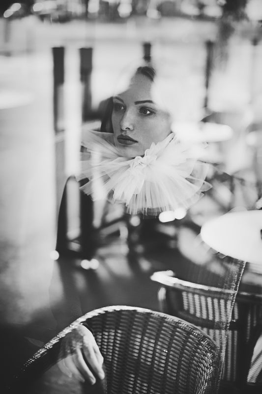 Cette photographie en noir et blanc de l’artiste Vanessa Moselle nous montre une jeune femme assise dans un bistrot. La femme est derrière une vitre ce qui donne des reflets à l’image. Elle regarde vers la gauche et se situe au centre de l’image. Au premier plan nous voyons des chaises en osier typiques des cafés parisiens.