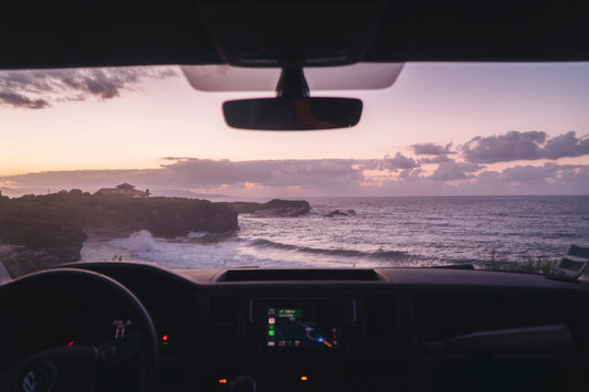 Photographie faites depuis le poste de conduite d’un van Volkswagen par le photographe Yorick Serriere en Cantabria en Espagne. Le van est garé en haut d’une falaise en bord de mer au coucher de soleil. Les couleurs du ciel et de l’eau sont dans les roses. Il y a une maison sur la falaise en face et des vagues. 