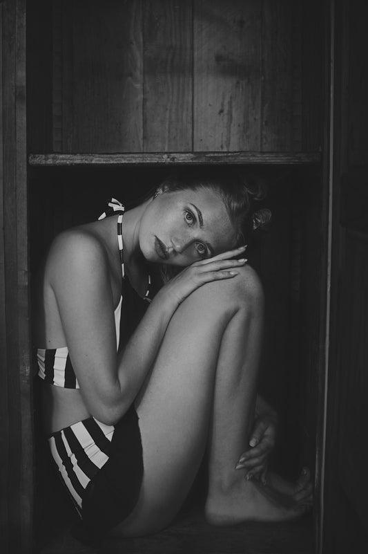 Photographie en noir et blanc de l’artiste Vanessa Moselle. Une femme en maillot de bain à rayures se trouve dans un boîte en bois et elle est contorsionnée comme pour se cacher dans un spectacle de cirque.