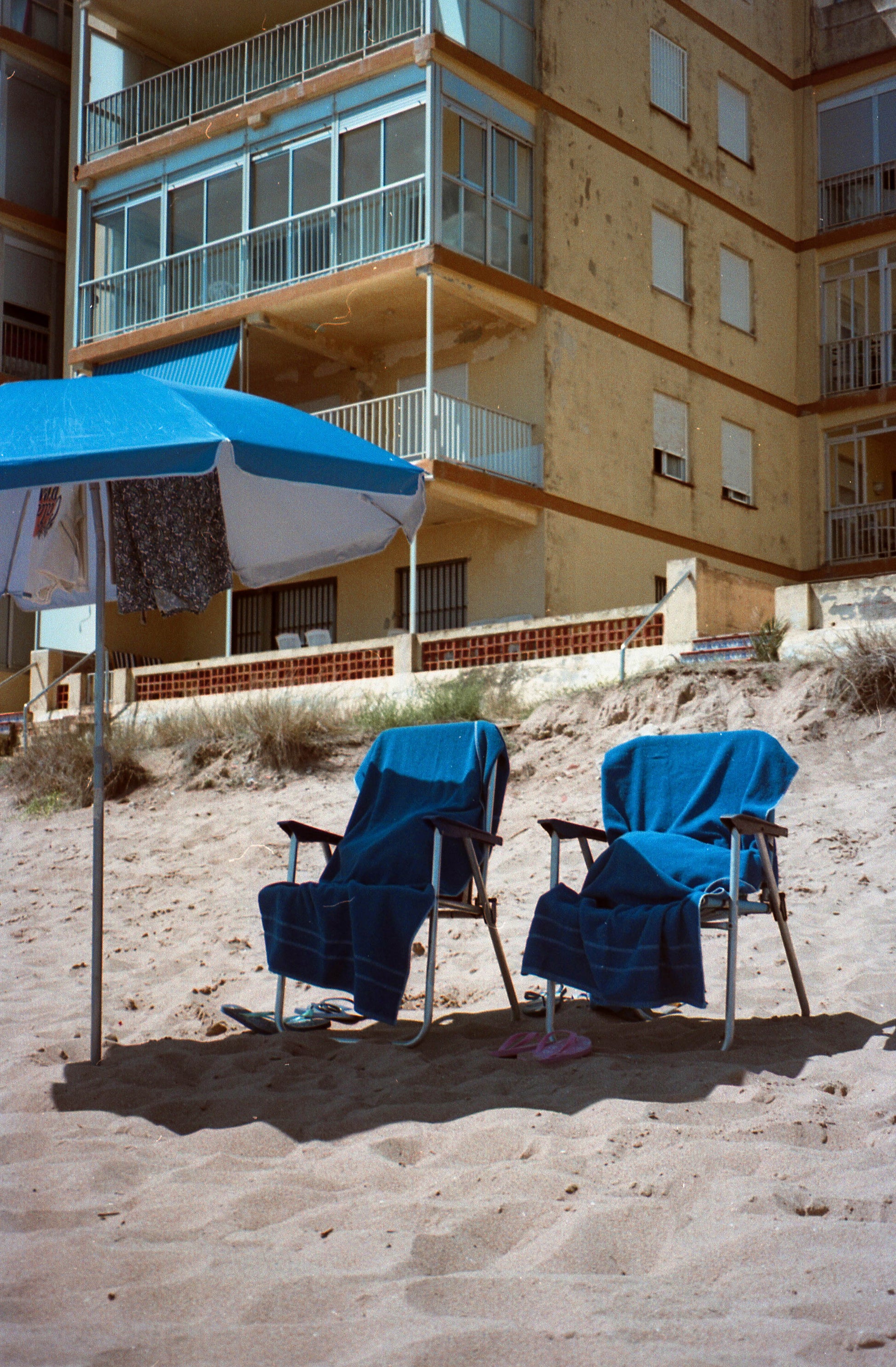 Photographie de deux fauteuils de plage vides. Ils sont recouverts d’une serviette bleue et sont à l’ombre d’un parasol lui aussi bleu. La plage est bordée d’immeuble anciens de couleur jaune avec des fenêtres bleues ciel. Photographie prise à Tavernes en Espagne par la photographe Alice Sevilla.