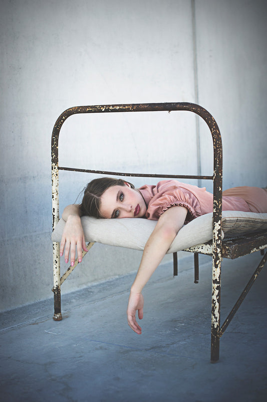 Photographie d’une femme allongée sur un vieux lit en métal. Cette photographie est prise par l’artiste Vanessa Moselle. La femme a les bras qui pendent dans le vide, elle est maquillée et porte un haut manches courtes de couleur rose pastel.