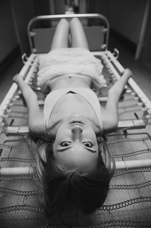 Photographie en noir et blanc d’une femme allongée sur un vieux lit d’hôpital réalisée par la photographe Vanessa Moselle. La femme sur la photo nous fixe du regard, elle est allongée à l’envers de nous. Elle porte un soutient gorge blanc et une jupe blanche. La photo est prise dans un long couloir.