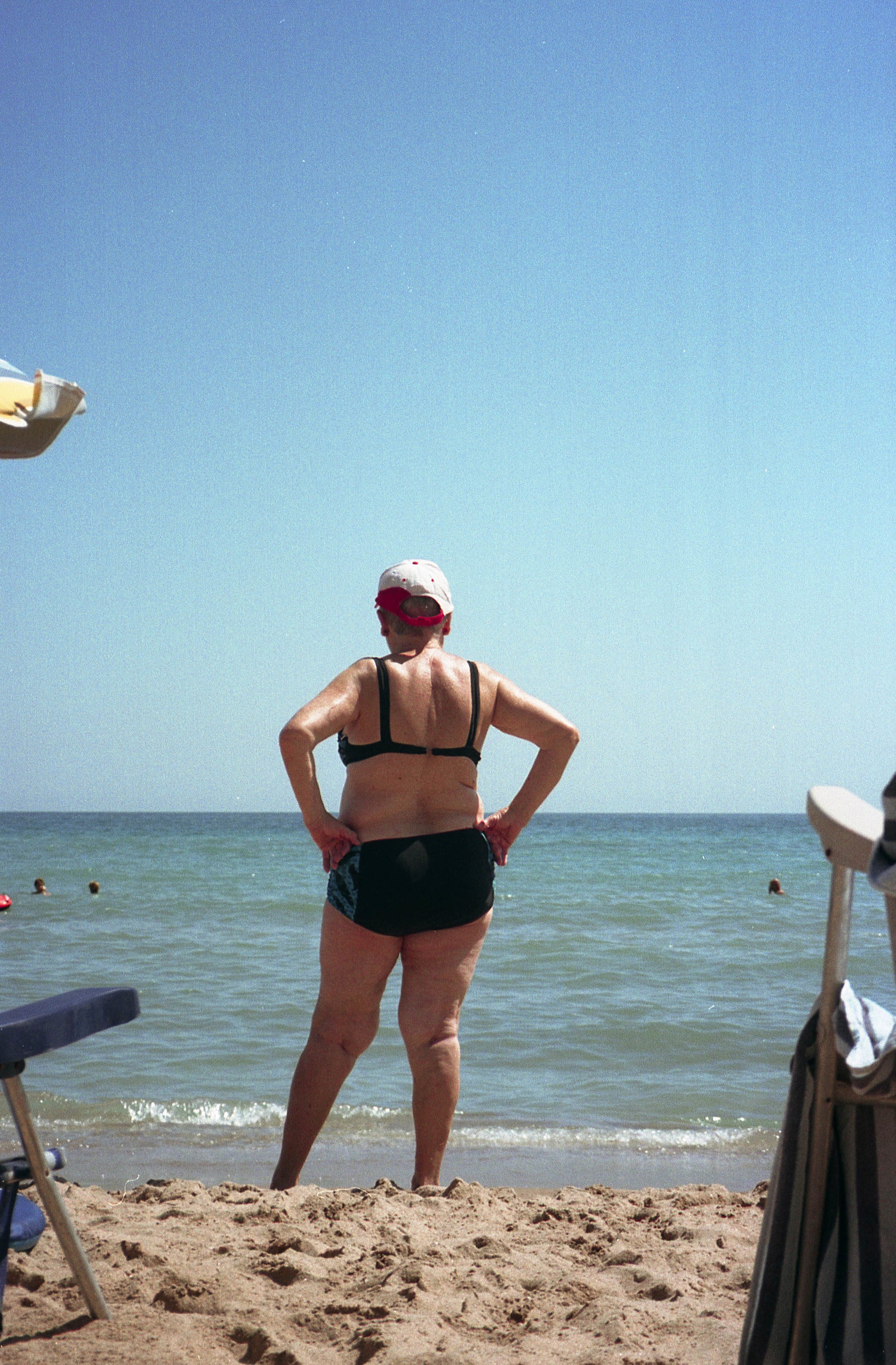 Photographie d’une dame âgée de dos regardant la mer. La femme est en maillot de bains deux pièces sur la plage de Tavernes en Espagne et pose ses mains sur ses hanches en regardant la mer. Nous avons l’impression qu’elle attends quelqu’un. Photographie prise par l’artiste Alice Sevilla.