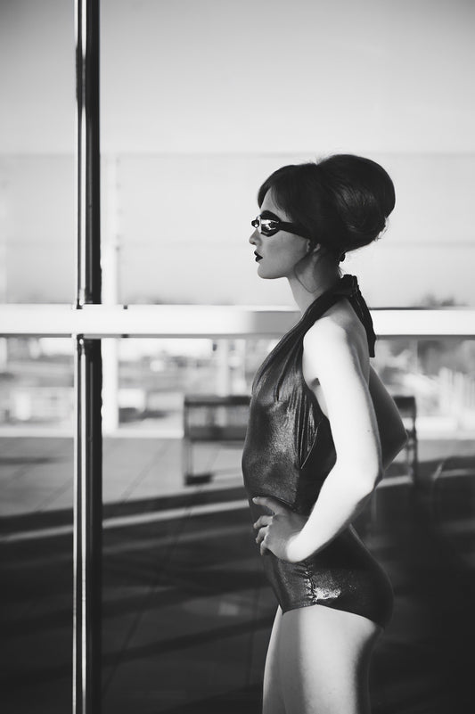 Une femme se tient debout face à une fenêtre sûrement d’un aéroport. Elle porte un maillot de bain une pièce et des lunettes de piscine. Ses cheveux sont attachés avec un chignon. Cette photographie en noir est blanc est réalisée par l’artiste Vanessa Moselle.