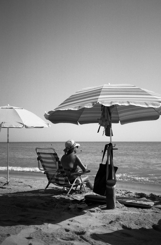 Une femme est assise sur une chaise pliante à la plage au bord de l’eau. Il y a un parasol mais elle n’est pas au soleil et elle regarde la mer comme si elle surveillait quelqu’un dans l’eau. Cette photo prise par Alice Sevilla à Tavernes en Espagne.