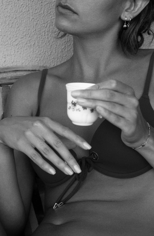 Photo en noir et blanc d’une femme en maillot de bains deux pièces qui prends son café. Son visage est coupé et la photo s’arrête au niveau de son ventre. Elle regarde sur le côté et nous laisse imaginer ce qu’elle regarde. Photo prise par l’artiste Alice Sevilla à Tavernes en Espagne.